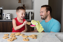 Happy Father And Son Feeding Glove Puppet In Kitchen While Eating Cookies