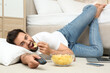 Lazy young man with bowl of chips watching TV while lying on floor at home
