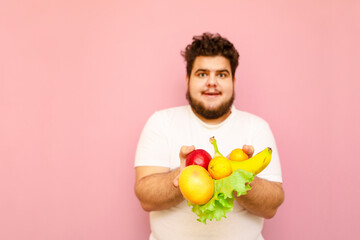 Wall Mural - Positive fat guy in white t-shirt isolated on pink background with healthy food, holds fruits and greens. Funny toast isolated on pink background with fruits, diet concept. Copy space