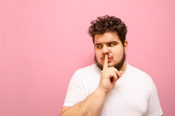 Wall Mural - Serious guy in a white T-shirt and overweight looks aside with a pensive face and shows a sign of silence. Concentrated fat man with a beard shows the gesture 