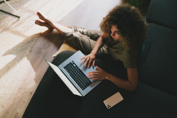 Home office concept. View from the top photo of a woman with long curly hair wearing cozy outfit working on a laptop while sitting on a floor with crossed legs. Remote work from the home office.