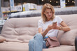 © mad_production - Cheerful mature woman using her smart phone, sitting on a couch at furniture store, copy space