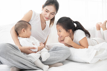 Asian mother and children girl baby laugh together at home. Asia mom holdind infant and play with sister sitting on white bed in living room at house family having fun together.