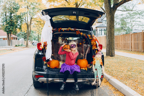 Trick or trunk. Child girl celebrating Halloween in trunk of car. Kid with red carved pumpkin celebrating traditional October holiday outdoors. Social distance during coronavirus covid-19.
