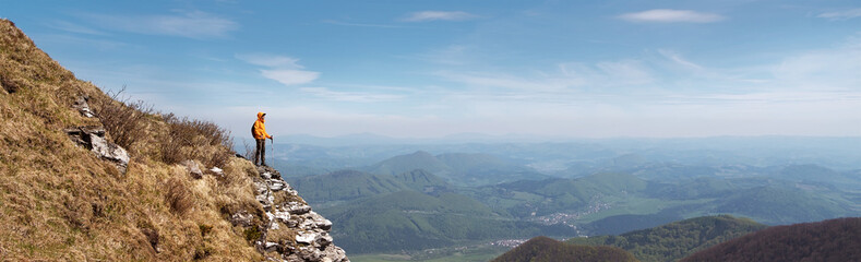 Dressed bright orange jacket backpacker with trekking poles staying on rocky cliff enjoying green valley at Mala Fatra mountain range,Slovakia. Active people and European tourism concept image.