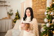 © Nelly - Beautiful girl in the living room at Christmas with a gift. Young woman in white sweater and white shawl for New Year's New Normal. Rustic interior with Christmas tree, sofa, gifts, hygge style.