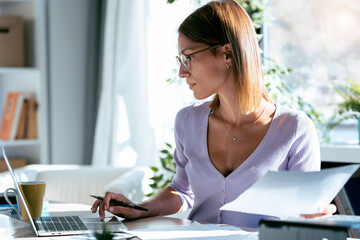 Concentrated young business woman working with computer while consulting some documents in the office at home.