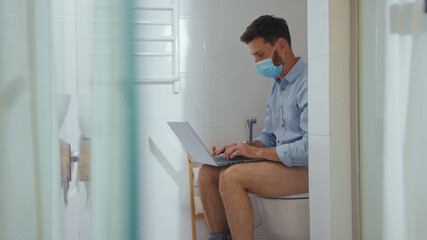 Happy businessman working on laptop wearing mask sit on the toilet at home restroom bearded funny wc isolation quarantine pandemic coronavirus covid-19 portrait close up slow motion