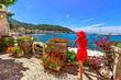 Back side of elegant woman in red in Marciana Marina, flowery old district Borgo al Cotone: meaning COTTON VILLAGE. Tourist on holiday travel, Italy Elba island. Harbor bay with cityscape background.