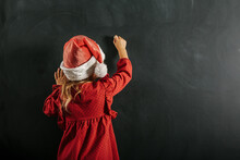 A Girl In Santa Claus Hat Draws Christmas Drawing On The Blackboard