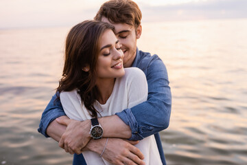 young man hugging brunette girlfriend with closed eyes beside sea at sunset