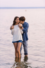 Wall Mural - Man hugging brunette girlfriend with closed eyes while standing in sea at sunset