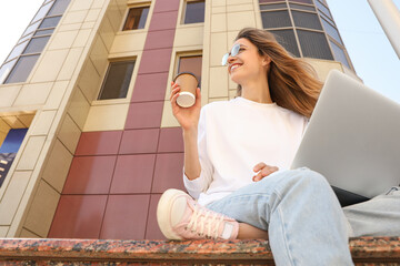 Wall Mural - Young woman with paper cup of coffee and laptop outdoors