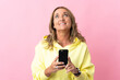 © luismolinero - Middle aged blonde woman over isolated pink background using mobile phone and looking up