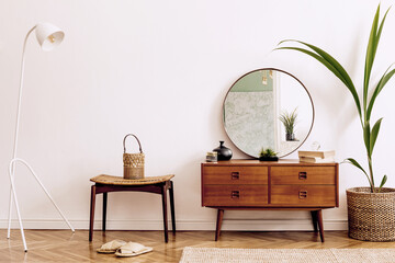 Minimalistic and stylish composition of living room with wooden commode, mirror, chair , lamp, tropicla plant in rattan basket and elegant personal accessories. Interior design, Home decor. template.