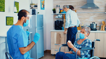 Disabled senior woman in wheelchair holding pills bottle from caregiver with face mask and visor during coronavirus pandemic. Social worker offering pills to handicapped elderly woman. Geriatrician