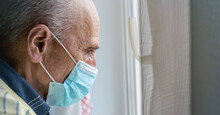Old Man In Disposable Face Mask Stands At White Window