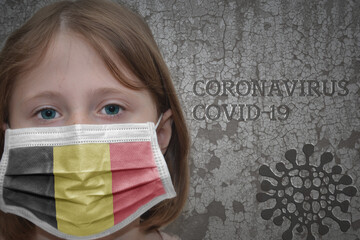 Wall Mural - Little girl in medical mask with flag of belgium stands near the old vintage wall with text coronavirus, covid, and virus picture. Stop virus concept