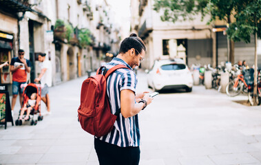 Rear view of young dark skinned hipster guy traveler walking on street with mobile phone browsing navigation app, young 20s male traveler exploring city using 4G connection on smartphone for searching