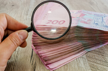 two hundred hryvnias are checked under a magnifying glass. Female hand with a magnifying glass and a stack of hryvnias. Ukrainian money on a wooden background.