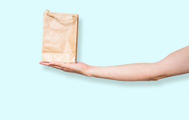 Wall Mural - Paper bag in hand on a blue pastel background. Concept of using ecological items. The man holds a brown bag with a gift, purchases in his hand. Giving gifts.