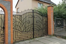 One Closed Gray Metal Gate With A Black Wrought Iron Pattern And Part Of A Brown Brick Fence Outside