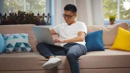 Concentrated young asian man wearing casual clothes sitting on couch using laptop computer