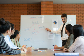 Young Caucasian manager in a tie shirt explaining on whiteboard about the new business plan to his asian business people team in the office meeting room.