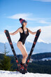 Woman in black swimsuit, hat and sun glasses, standing with skis under sun at ski resort. Blue sky, forests, mountains on the background. Ski season and winter sports concept