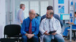 © DC Studio - Doctor explaining treatment to senior man in hospital waiting area. Woman with walking frame arriving at medical examination.