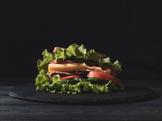 Sandwich with lettuce, tomatoes, cheese, bacon, cucumbers on a stone plate on a dark wooden background. Vintage style.