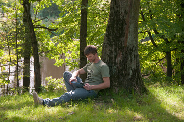 Man sitting under the tree by the lake and using his digital tablet. Guy working outdoors.