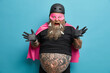 © WHstudio Leushin N - Emotional bearded European man looks excited and cheerful raises hands keeps mouth opened wears pink mask cape helmet poses against blue background. Male superhero with thick beard fat abdomen