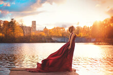 Romantic Portrait Of A Young Girl In A Long Red Dress Standing In The Wind Against The Background Of An Ancient Castle And Autumn Nature