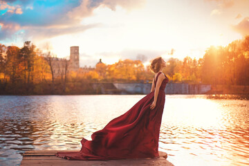 Romantic portrait of a young girl in a long red dress standing in the wind against the background of an ancient castle and autumn nature