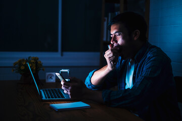 Asian businessman working late night feeling tired, sleepy yawning stressful in front using computer laptop smart mobile phone technology, overtime busy schedule hard work at home modern office space