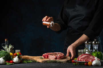 The chef in black apron pours pepper on fresh meat steak on wooden chopping board at the professional restaurant or hotel kitchen. Cooking tasty healthy meal. Food concept. Delicious dish.