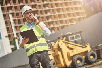 Wall Mural - Important call. Young civil engineer or construction supervisor wearing helmet talking by phone while inspecting building site