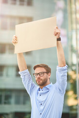 Portrait of a young male activist wearing blue shirt and eyeglasses holding empty sign board while standing outdoors, vertical shot