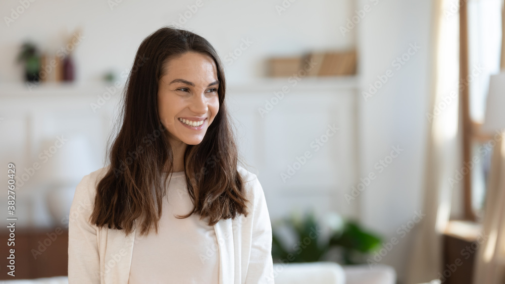 Happy confident young female owner, buyer or tenant of new flat standing in living room alone and looking aside with cheerful white smile feeling independent and free resting at home, copy space - obrazy, fototapety, plakaty 