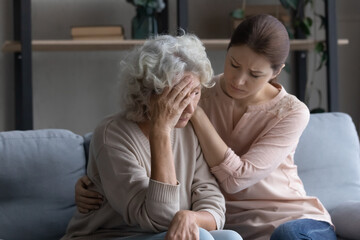 Wall Mural - Caring grownup daughter comforting frustrated unhappy mature woman, hugging, sitting on couch at home, upset elderly female feeling unwell, touching forehead, suffering from migraine or head ache