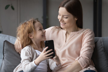 Smiling cute little girl and young mother having fun with phone, sitting on cozy couch at home, happy family enjoying leisure time together, shopping online, watching video, browsing apps