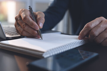 Close up of man hand writing on paper notebook with a pen while working on laptop computer