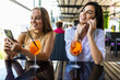 © F8  \ Suport Ukraine - Positive young women recreat together at cozy restaurant, use phones, enjoy tasty cocktails.