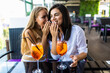 © F8  \ Suport Ukraine - Two women sharing secrets and covering faces with hands while drink cocktails in cafe.