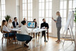 © fizkes - Confident female speaker or coach stand make whiteboard presentation at meeting with diverse employees. Successful woman trainer talk present business project on flip chart for workers in office.