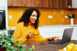 © Carlos David - Portrait of a mid woman with black curly hair smiling while using laptop computer in kitchen