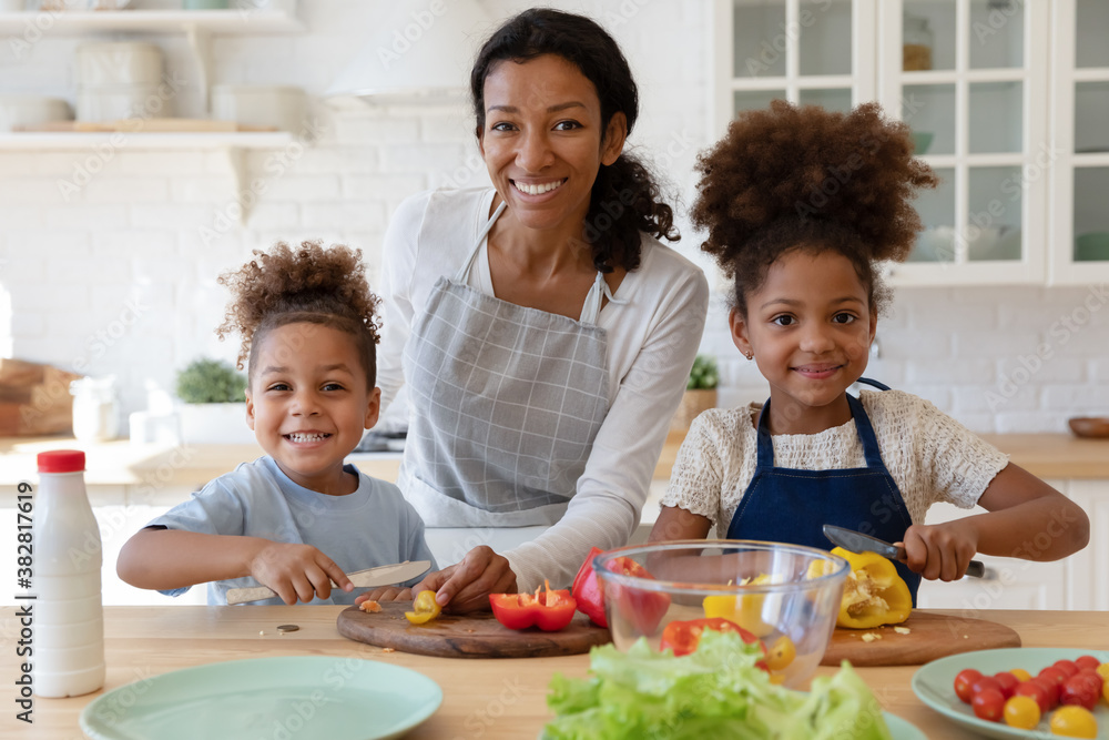 Portrait of happy young african american mother enjoying cooking healthy vegetarian food together with joyful little cute children in modern kitchen, smiling adorable family looking at camera. - obrazy, fototapety, plakaty 