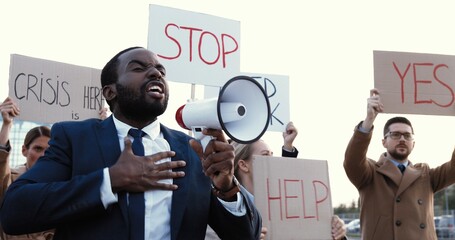 African American handsome man in suit and tie talking in megaphone outdoor at protest against unemployment in multi ethnic crowd. Male having speech at strike against inequality and for human rights.