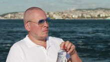 Video, An Adult Man Drinks Water From A Plastic Bottle On The Background Of Turquoise Sea Water, On The Beach On A Sunny Day In Summer. Ships Float On The Water Out Of Focus.